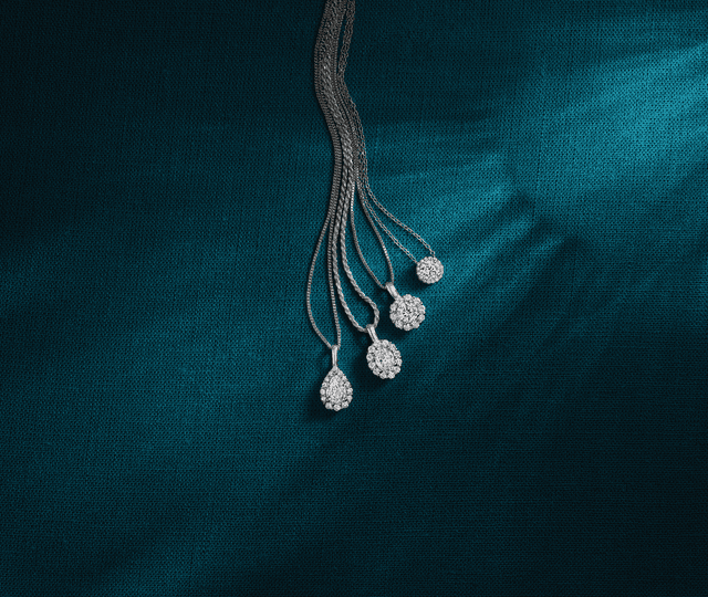 Collection of Motek Jewelry sparkling diamond halo and teardrop pendant necklaces layered over a textured teal-blue fabric.