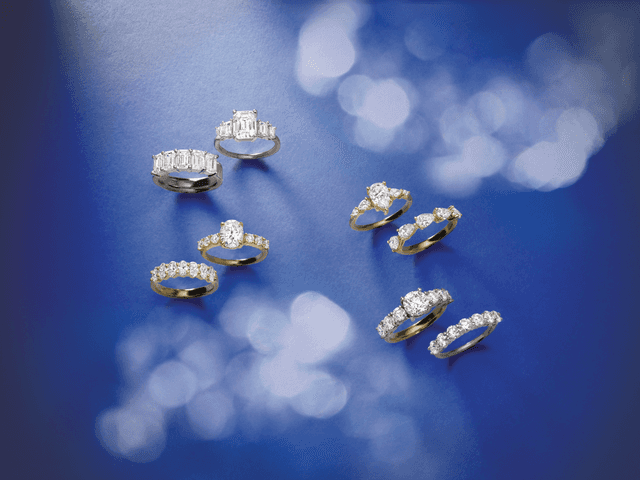 Collection of Motek Jewelry diamond engagement and eternity rings arranged across a deep royal blue background with soft bokeh light.