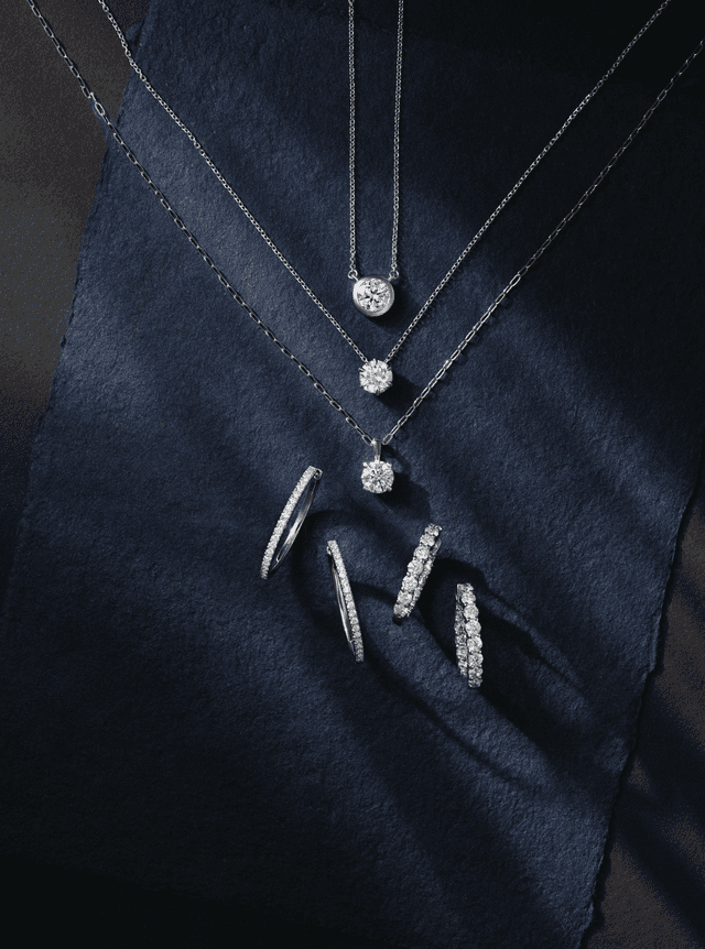 Motek Jewelry diamond solitaire pendant necklaces and diamond hoop earrings displayed against a rich navy silk textile.