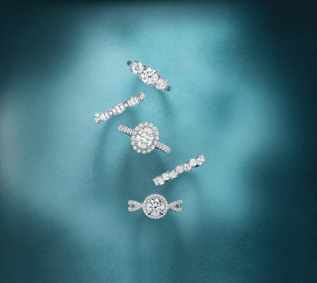 Sparkly Motek Jewelry oval and round halo diamond engagement rings paired with eternity bands over soft aqua velvet fabric.