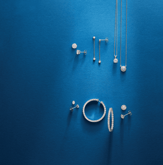 Motek Jewelry diamond pendant necklaces, hoop earrings, bangles, and bezel studs arranged on a textured deep navy surface.