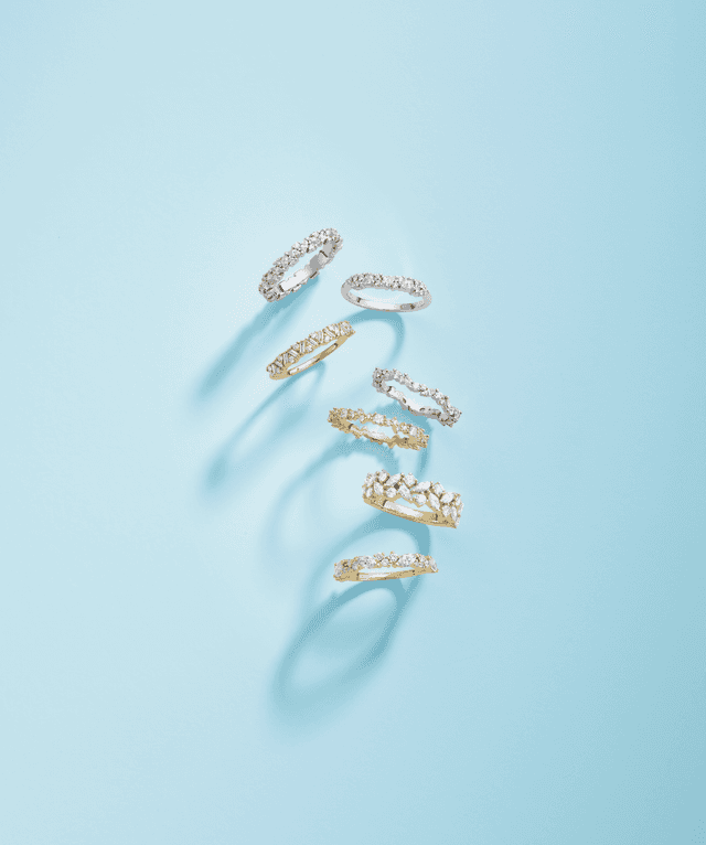 Variety of Motek Jewelry pavé, bezel, and marquise-cut diamond eternity bands floating over a bright pastel blue background.